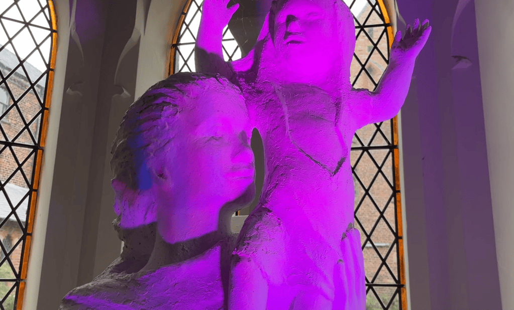 Sculpture of woman holding a baby high on her shoulder. Lit in bright magenta light.